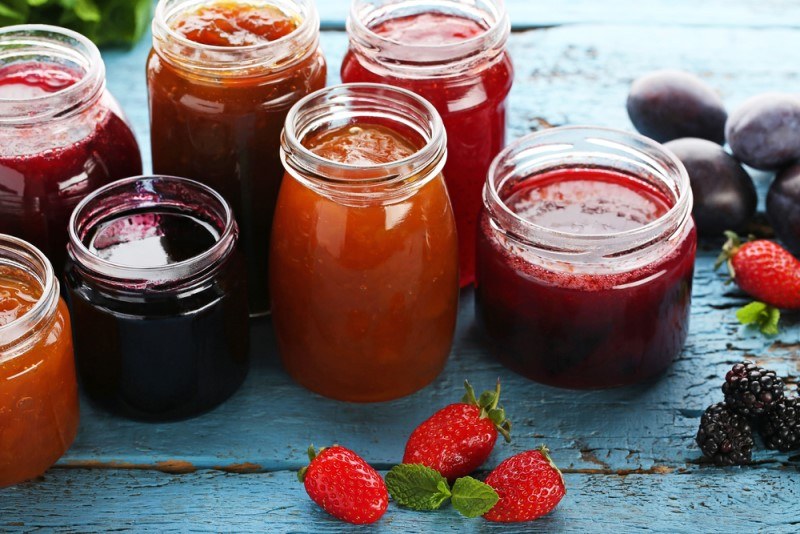 A selection of various jams in open glass jars rest on a wooden surface near strawberries, blackberries, and plums.