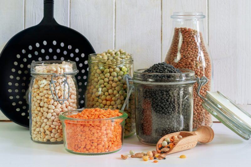 different jars filled with different legumes, with a black colander at the back and a wooden scoop with legumes at the foreground