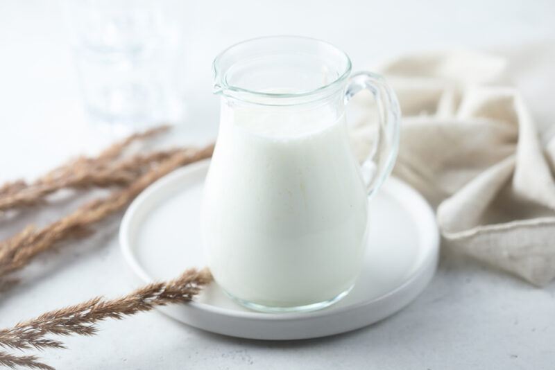 on a white concrete surface is a clear jug of cream resting on top of a white round dish, a light beige table cloth and brown dried stalks beside it