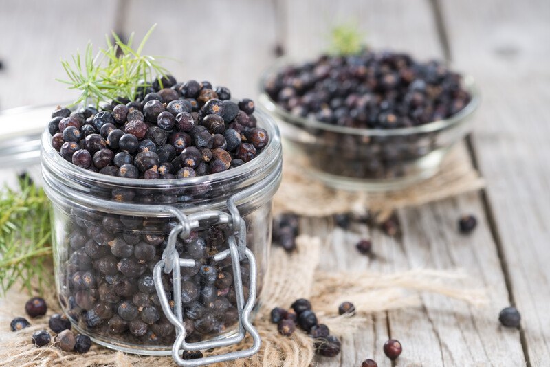 on a wooden surface is a glass jar full of juniper berries resting on a burlap sack with a small glass bowl full of juniper berries also resting on a burlap sack at the back