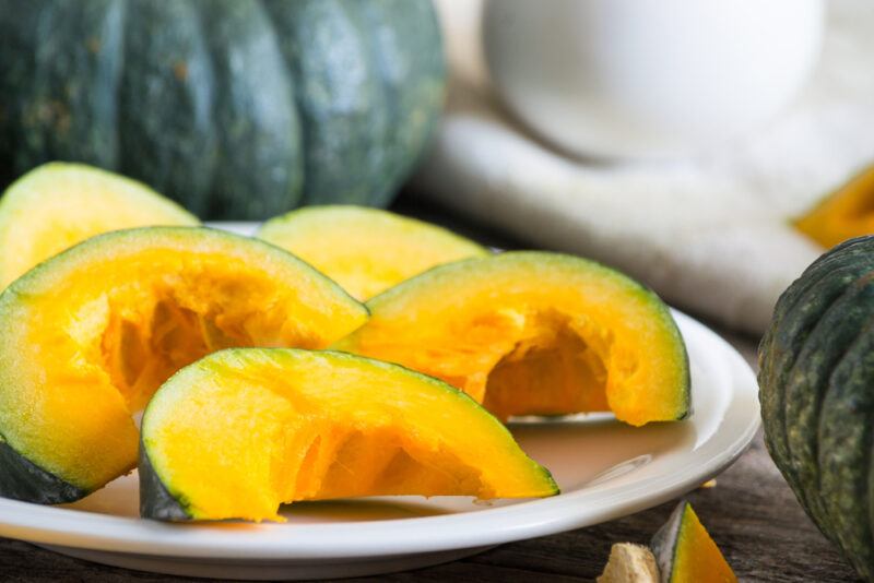 closeup image of sliced kabocha on a white ceramic dish with sliced kabocha, at the back is a whole kabocha with a small white ceramic jar resting on a white table napkin