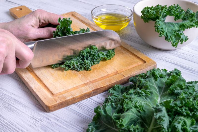 on a white wooden surface is a woman's hands chopping kale on a wooden chopping board with kale leaves beside it and white bowl full with kale and bowl of olive oil at the back