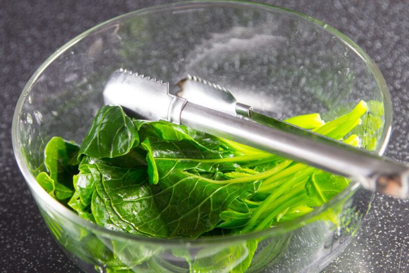 closeup image of a clear glass bowl with boiled kale, a type of dark leafy green vegetable, and a metal tong