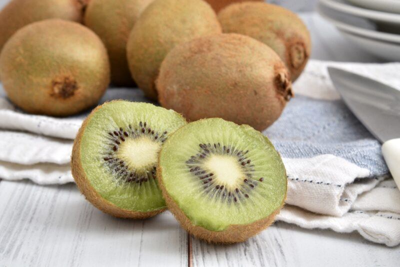 on a white wooden surface is a halve kiwi with a spread of whole kiwis at the back resting on a white and blue table napkin