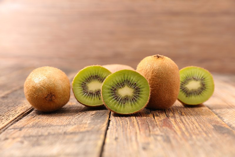 Three uncut kiwi fruit and three cut kiwi halves rest on a wooden table.