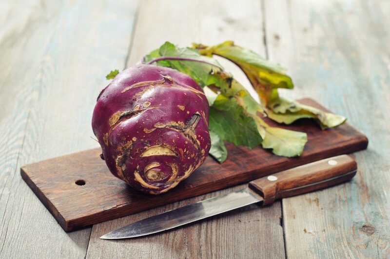 on a wooden surface is a whole kohlrabi resting on a small wooden chopping board with a knife beside it