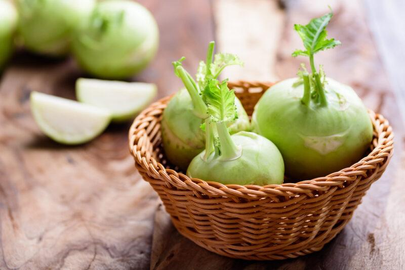 on a wooden surface is a weave basket with three whole kohlrabies with a couple of whole kohlrabies and cut kohlrabi at the back