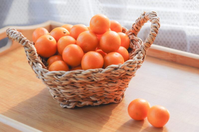 on a wooden tray is a weaved basket with handles full of kumquats with a couple of loose kumquats beside it