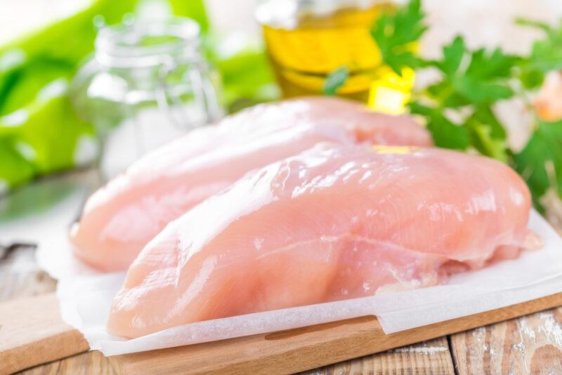 closeup image of a couple of skinless chicken breast resting on a piece of white paper on a wooden chopping board, at the back are herbs and a jar of olive oil