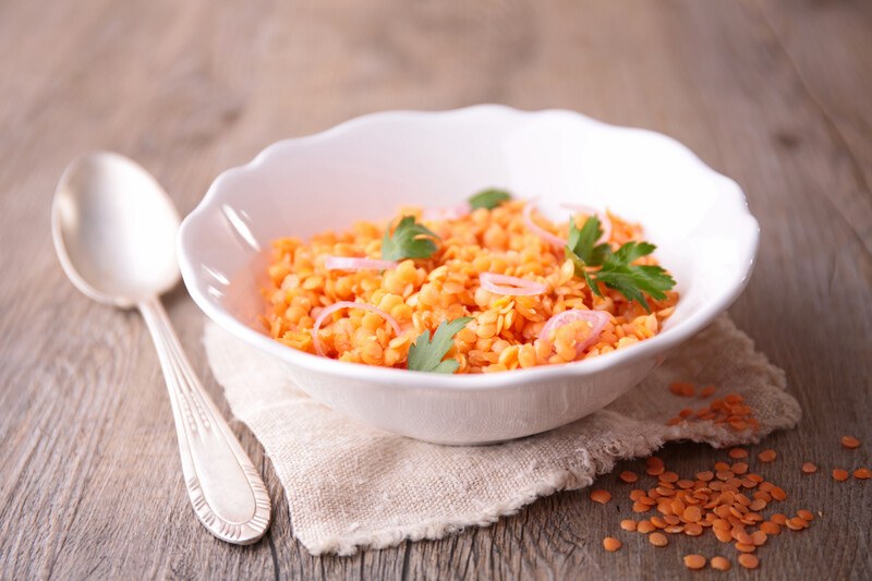 A white porcelain bowl filled with orange lentils rests on a cloth near a spoon on a wooden tabletop.