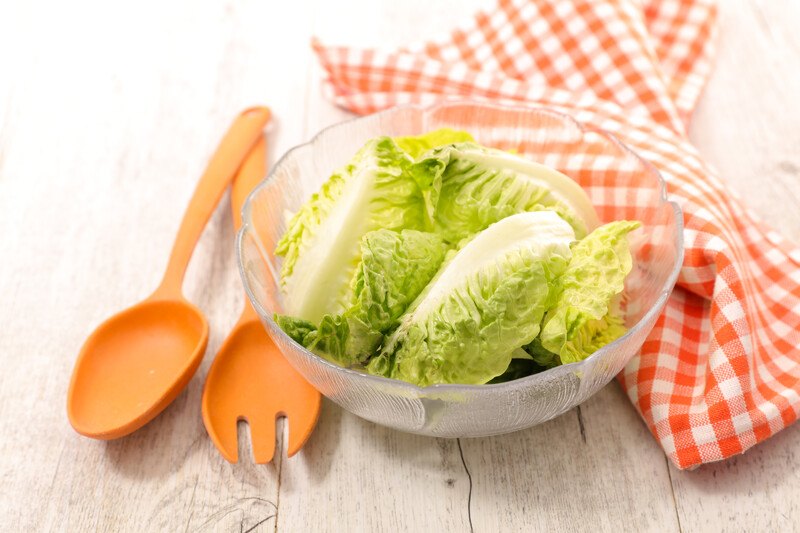 on a wooden surface with a clear bowl of lettuce with wooden spoon and fork beside it and a red and white checkered table napkin