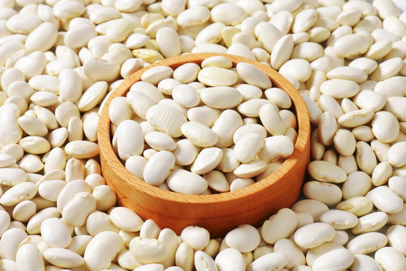 A wooden bowl of white raw lima beans is surrounded by more raw white lima beans.