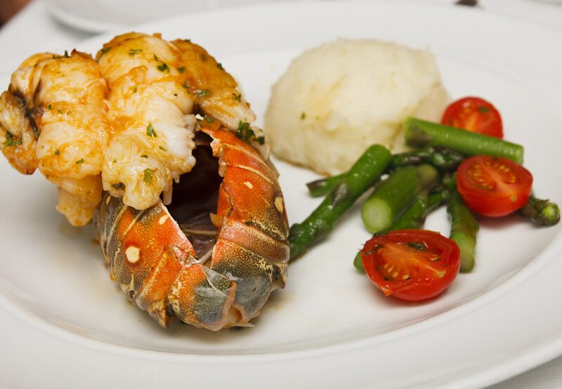 This photo shows a cooked lobster tail on a plate with mashed potatoes, asparagus, and grape tomatoes.