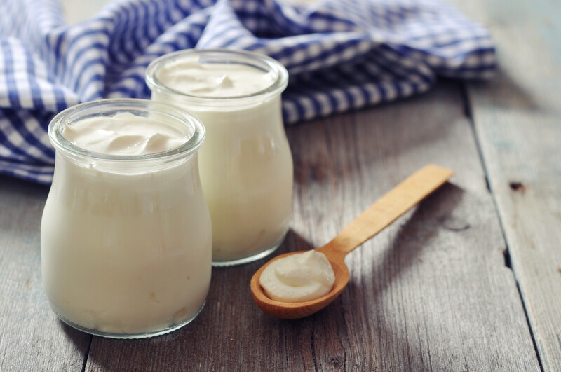 on a wooden surface is a couple of jar with low fat yogurt and wooden spoon with yogurt beside it, with blue and white table napkin