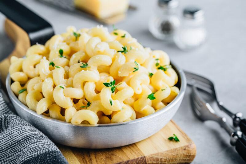 on a concrete surface is a bowl full of macaroni and cheese resting on top of a wooden chopping board with a grey table napkin beside it, beside the chopping board is a couple of forks, behind it a salt and pepper shaker