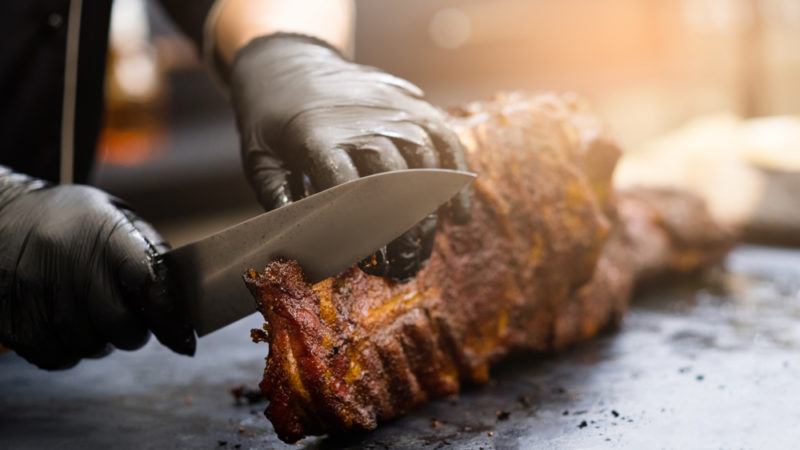 man with black gloves cutting into smoked ribs with a sharp knife