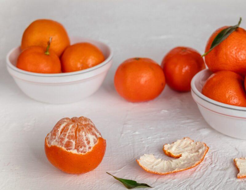 on a white surface is a couple of white bowls with mandarins around are loose mandarins with one partially peeled