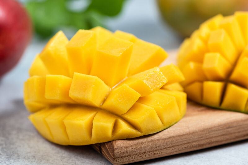 a closeup image of sliced mangoes resting on a wooden surface, at the back is some greens and red fruit