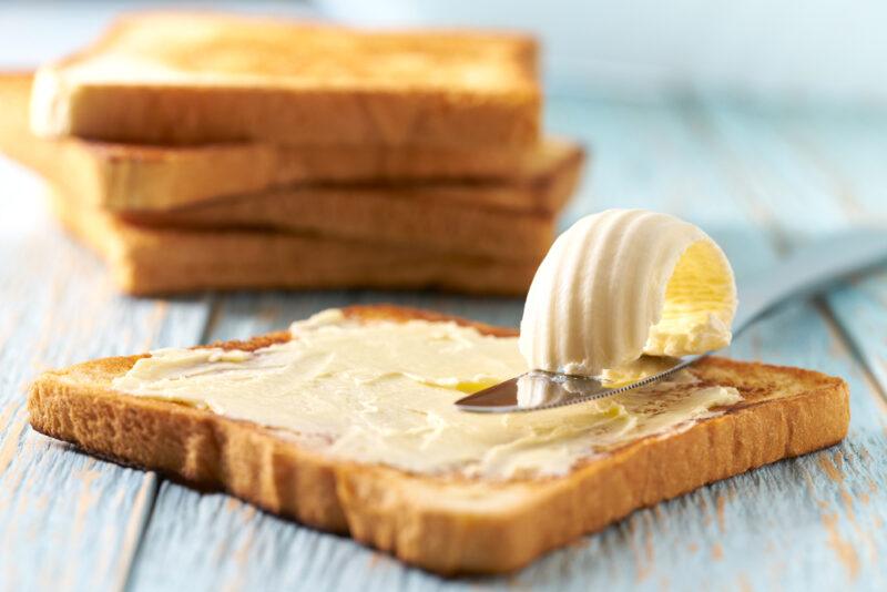 on a rustic looking bluish wooden surface is a stack of toasted white bread slices, one with a spread of margarine