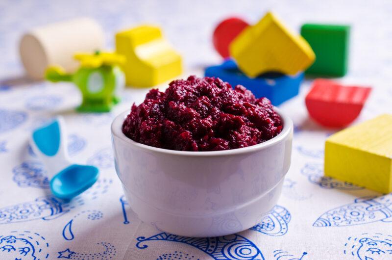 white bowl full of mashed red beets with a blue plastic spoon for baby, at the back are brightly colored blocks for babies