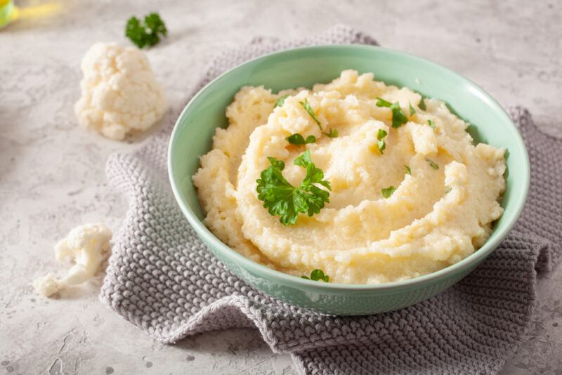 on a concrete surface is a light green bowl with mashed cauliflower with herbs on top, resting on a grey table napkin with a couple of cauliflower florets beside it