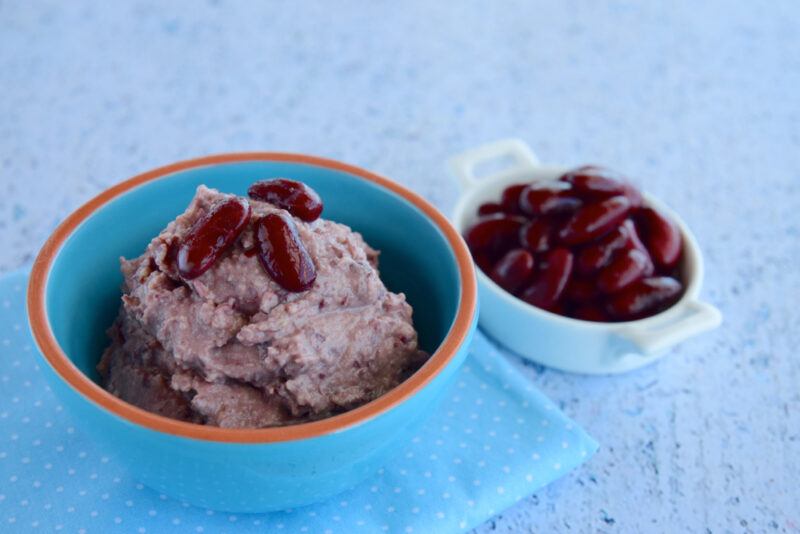 on a marble surface is a blue bowl with orange rim full of mashed kidney beans with whole kidney beans on top, resting on top of a light blue table napkin with white dots, beside it is a small white bowl full of kidney beans