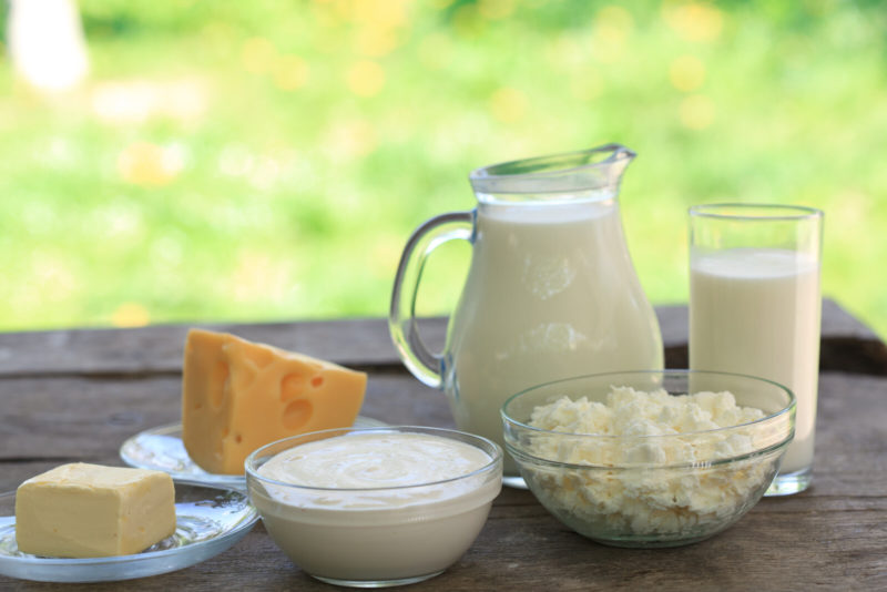 a pitcher and a glass of milk, fresh yogurt, and three different types of cheese on a wooden table.