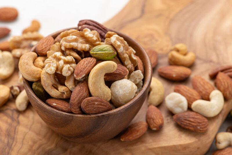 a wooden bowl of mixed nuts resting on a wooden board with loose mixed nuts around it