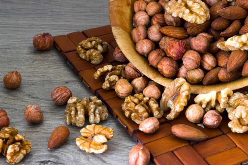 on a wooden surface is a closeup image of almonds, walnuts, and hazelnuts pouring out of a brown wooden bowl resting on top of a brown wooden placemat 