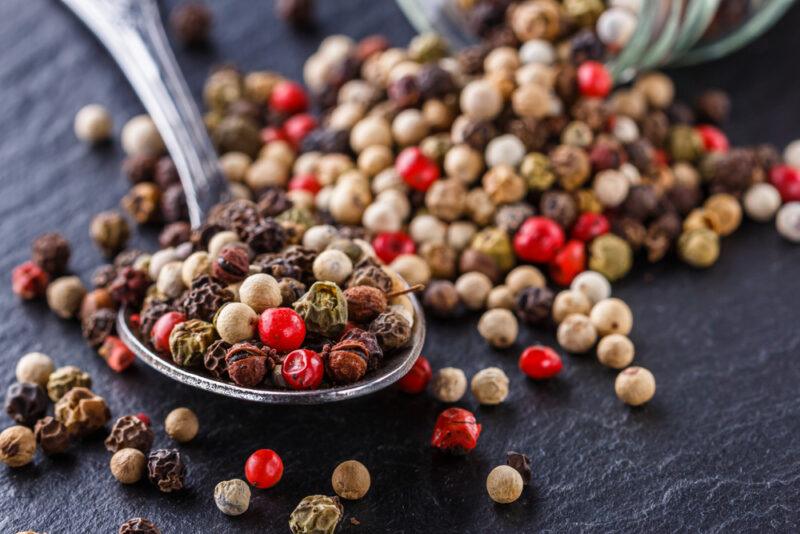 on a dark surface is a closeup shot of mixed peppercorns pouring out of a small jar with a silver spoon full of mixed peppercorns as well