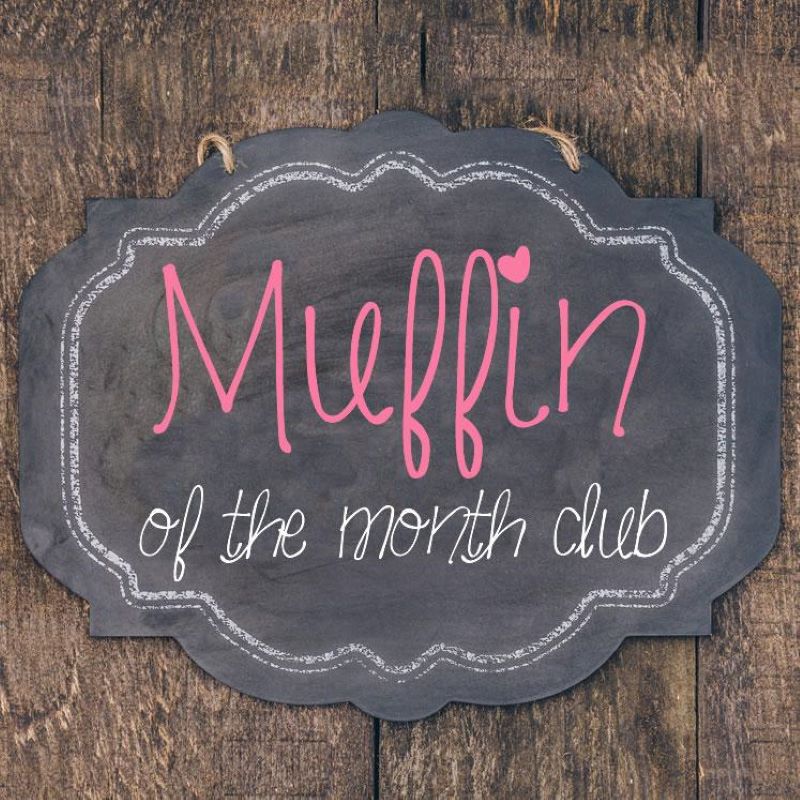 black chalk board sign with white outline and pink writing that says muffin and below in white of the month club sitting on a wooden plank background