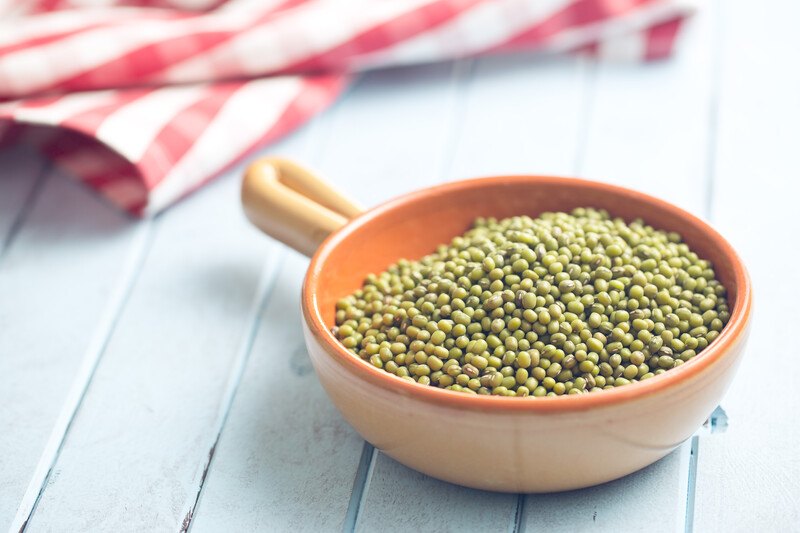 on a white wooden surface is a brown dish with handle full of mung beans, behind it is a white and red table napkin