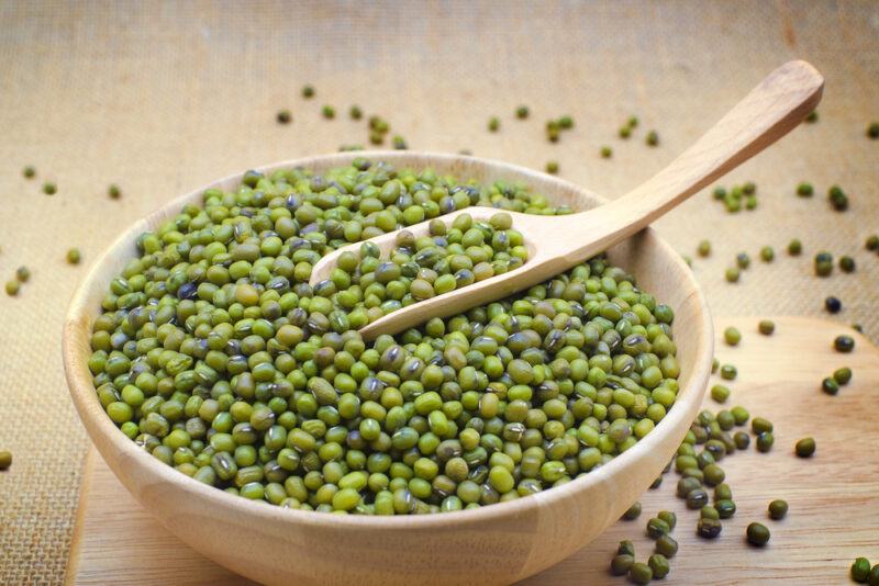 on a wooden board is a wooden bowl with mung beans, with a wooden spoon shoved in it, around it are loose mung beans