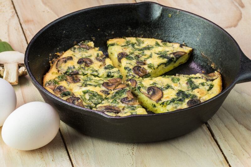 on a wooden surface is an iron skillet pan with mushroom and spinach frittata with a couple of eggs and sliced mushrooms beside it