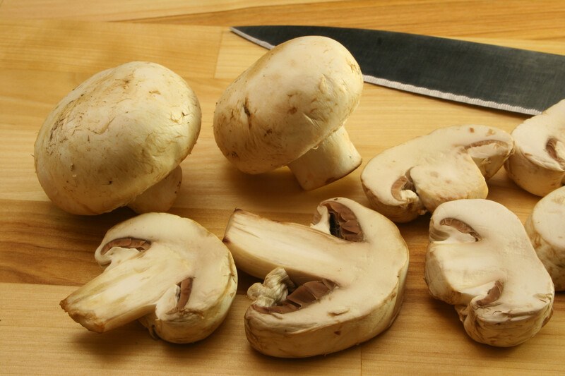 Two whole brown mushrooms and several mushroom slices lie on a cutting board next to a knife.