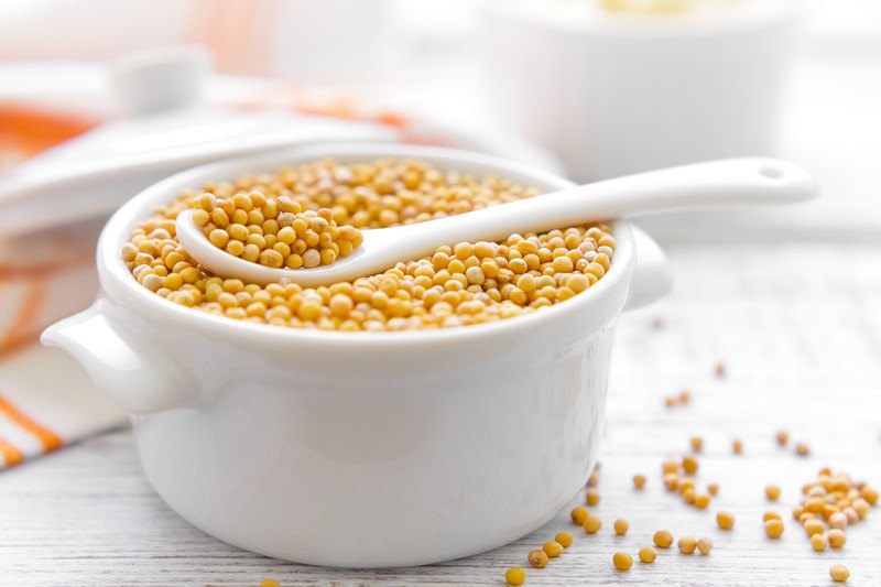 mustard seeds in a small white ceramic container with a tiny ceramic spoon, resting on a white-colored wooden surface with loose mustard seeds around it