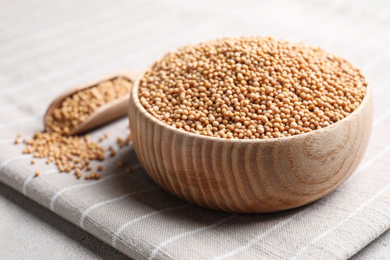 resting on a grey table napkin with white stripes is a wooden bowl of mustard seeds with a wooden scoop beside it with mustard seeds as well, with loose mustard seeds around it