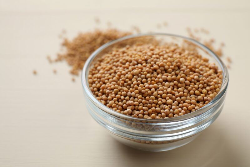 on a beige-colored surface is a clear glass bowl with mustard seeds, behind it is a small pile of ground mustard seeds