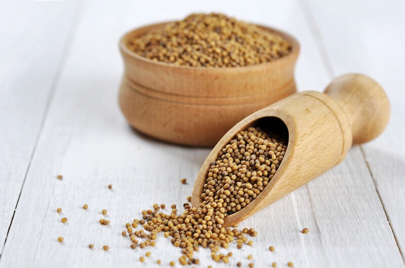 on a white wooden surface is a wooden bowl full of mustard seeds with a wooden scoop with mustard seeds beside it