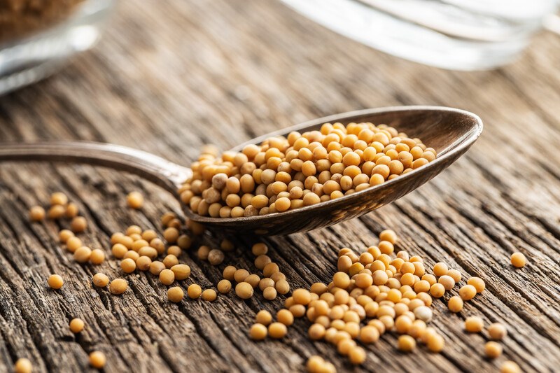 on a rustic wooden surface is a silver spoon with mustard seeds, with loose mustard seeds around it