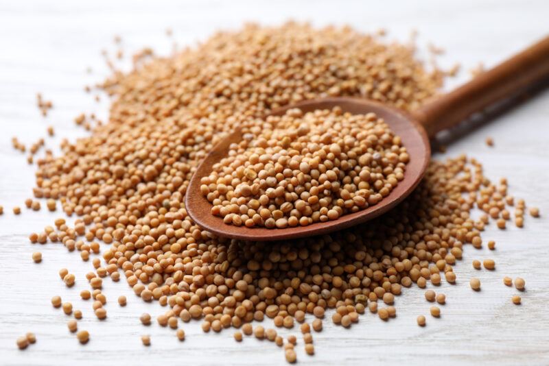 on a white wooden surface is a close up image of a mound of mustard seeds with a wooden spoon on top also with mustard seeds