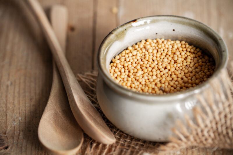 on a wooden surface is a ceramic tan-colored little pot with mustard seeds, resting on top of a burlap with a couple of wooden spoons beside it