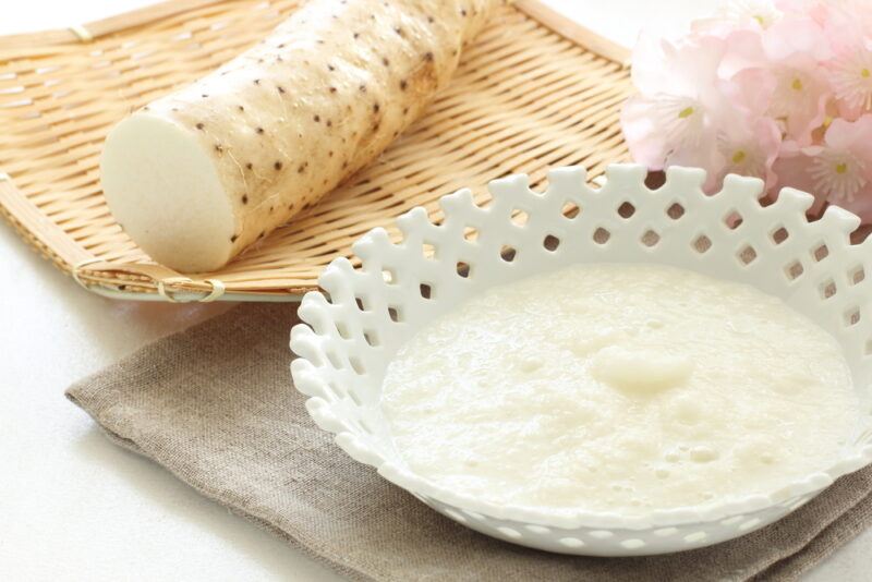 on a white surface is a nagaimo on a shallow weaved tray with a white bowl with grated nagaimo resting on a brown table napkin