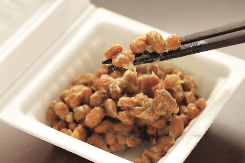 This photo shows two black chopsticks with natto on them hovering above an open white container of brown natto.