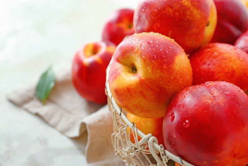 a closeup shot of nectarines on a weaved white metal basket, resting on top of a brown kitchen towel with a couple of nectarines and a leaf at the back