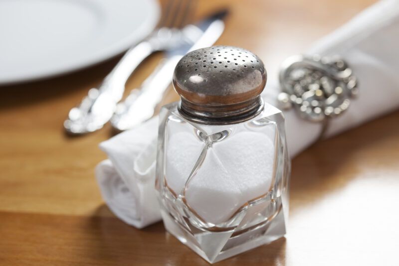 on a wooden surface is a vintage salt shaker with table napkin, silver ware, and partial image of a white plate at the back