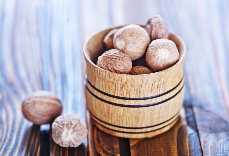 on a wooden surface is a closeup image of a wooden container full of whole nutmeg, beside it is a couple of nutmeg