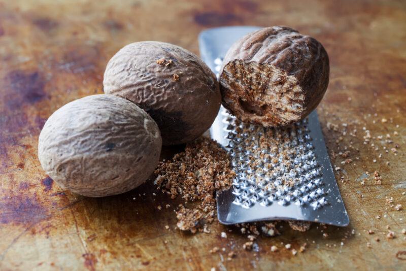 on a rustic looking wooden surface are two whole nutmegs and one partly grated seating on a metal grater 