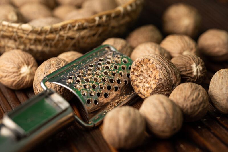 on a dark wooden surface is a closeup image of nutmegs with a metal grater, at the back is a weaved basket with nutmegs as well