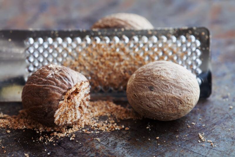 on a rustic looking concrete surface is a closeup image of a metal grater with a few nutmegs, one partially grated